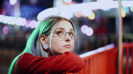 Hipster girl with blue dyed hair, golden sequins as freckles. Woman with nose piercing, transparent glasses, ears tunnels, unusual hairstyle stands in amusement night parkの写真素材