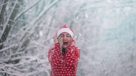 Cheerful woman enjoys winter wonderland. Snow flies on her and she is happy, rejoices cold weather. Girl in red Christmas sweater and Santa hat.の写真素材