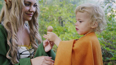 Fairy elf cosplay woman treats little baby boy hobbit with candy in green forest. Halloween concept, fairytale characters, kids.の写真素材