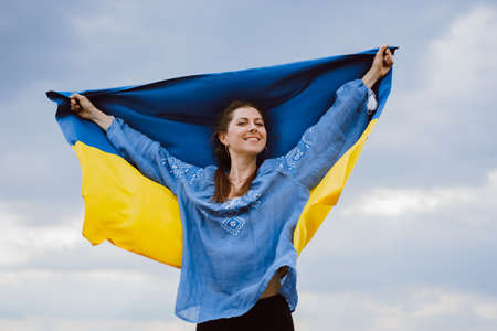 Happy free ukrainian woman with national flag on dramatic sky background. Portrait of lady in blue embroidery vyshyvanka shirt. Ukraine, independence, patriot symbolの写真素材