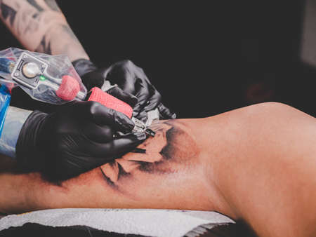 Close up view of tattoo artist demonstrates the process of getting black skull tattoo with paint. Concentrated female coloring contour ink on leg with gun. Neon light.の写真素材