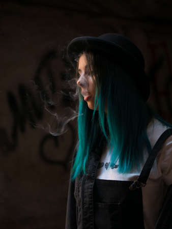Emo girl smoking cigarette.Street punk or hipster woman with blue colorful dyed hair, hat, piercing,lenses,ears tunnels and unusual hairstyle stands in backyard. beautiful smoke.の写真素材
