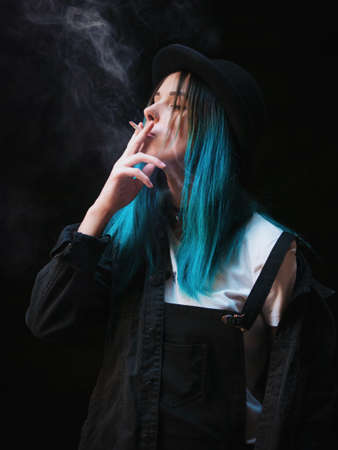 Emo girl smoking cigarette.Street punk or hipster woman with blue colorful dyed hair, hat,...