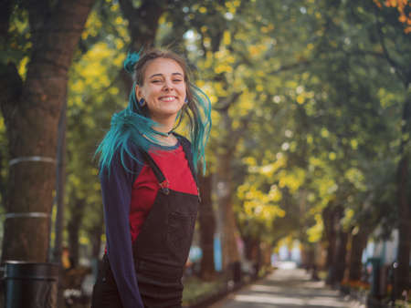 Street punk or hipster girl enjoying empty old European park. Portrait of teen girl with blue dyed hair,piercing in nose,violet lenses and unusual hairstyle dancing.Carefree concept.の写真素材