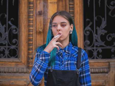 Young punk girl smoking cigarette by the wooden door.Hipster with blue dyed hair. Woman with piercing in nose, violet lenses, ears tunnels and unusual hairstyle.の写真素材