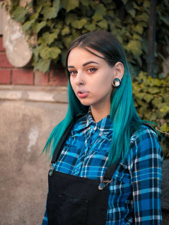 Young attractive punk girl smoking cigarette. Hipster with blue unusial dyed hair, piercing in nose, violet lenses, ears tunnels on empty European old street.の写真素材