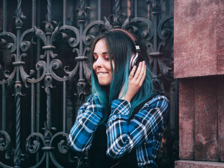 Street hipster girl listening music with brown headphones. Teen girl with blue dyed hair,piercing in nose,violet lenses,unusual hairstyle sitting near historic building.の写真素材