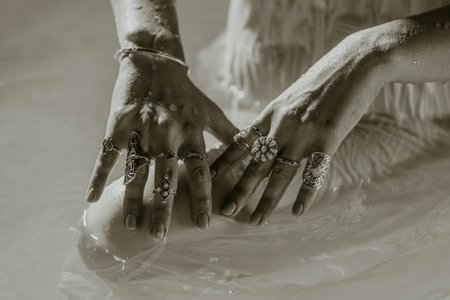 Beautiful gypsy woman demonstrates boho jewelry, rings with turquoise stones on hands. Black and white. Girl in white dress showing accessories in blue water. Femininity, trend, hippie style concept.の写真素材