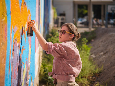 Beautiful Young Girl making a colorful graffiti with aerosol spray on urban street wall. Cinematic toned shot. Creative art. Talented woman in glasses drawing picture.の写真素材