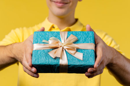 Man gives gift box by hands to camera on yellow background. Guy smiling, he is happy with present. Offer surprise for youの写真素材