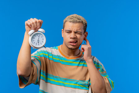 Woken up by alarm clock sleepy man holding it in hand. Blue background. Early 6 oclock in morning. Lazy guy didnt get enough sleep, concept of passing time.の写真素材