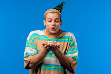 Happy birthday man making wish - blowing candle on cake. Curly haired guy laughs, celebrating...