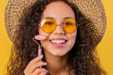 Curly woman doing face massage with rose quartz stone roller yellow backgroundの写真素材