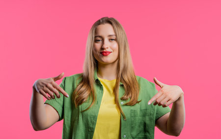 Blonde woman pointing down to advertising area. pink background. Young blogger asking to click to subscribe below. Copy space for your commercial idea, promotional content. high qualityの写真素材