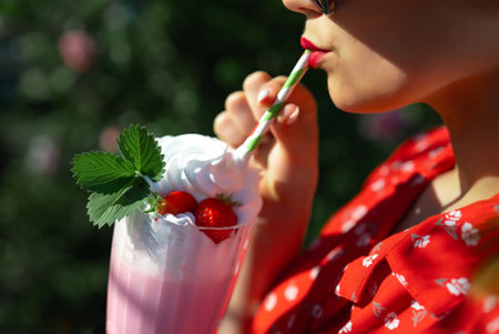 Pretty woman drinking strawberry milkshake cocktail with straw, nature backdrop