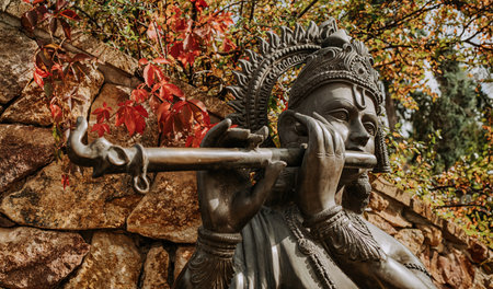 Bronze statue of Lord Krishna playing on flute. Autumn nature. Hinduism religion