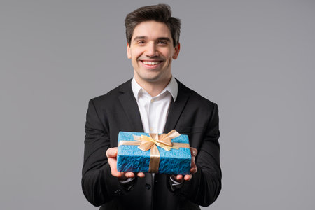 Handsome Businessman In Suit Gives Gift Box Present To Camera. Man is happy, smiling on grey office wall background. Positive mood, holiday, bonus, birthday concept.の写真素材