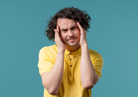 Young man having hypertension headache, blue studio. Guy putting hands on head