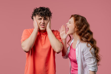 Grumpy woman quarrels on husband or boyfriend On pink. Tired man covers earsの写真素材