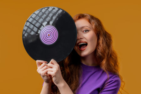 Positive red haired woman with vinyl record disc on yellow. Hobby, music lover, collection,...