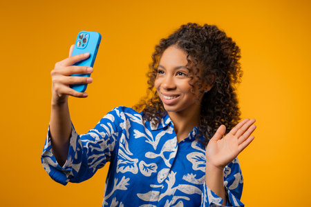 Happy african american teenager girl talking camera as video call, makes selfie or record vlog as bloggers on yellow studio wall background.の写真素材