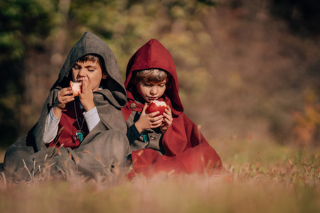 Hobbits boys in costumes sitting on autumn ground to eat apples. Fantasy journey, halloween, fellowship cosplayの写真素材