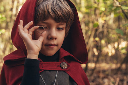 Little cosplayer hobbit boy in cape costume in autumn forest admiring golden ring. Fantasy journey, halloween, fellowship cosplayの写真素材