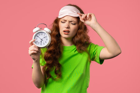 Woken up by alarm clock sleepy young woman holding it in hand. pink backgroundの写真素材