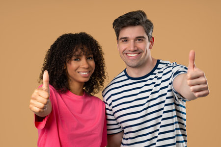Positive woman of color and white man give like, thumbs up gesture. Couple on beige
