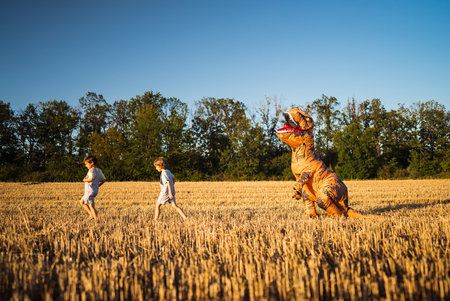 Little kids boys chased by person in inflatable dinosaur T-Rex costume in fieldの写真素材