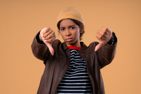 African Teen Girl With Dislike Signs. Stylish Lady Showing Thumbs-Down expressing discontent on beige studio background.