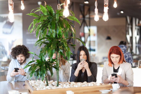 Four friends sitting at a cafe bar looking at phones.の写真素材