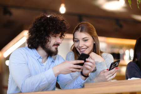 Two friends sitting at a cafe bar looking at phone. Positive and happy face expressions.