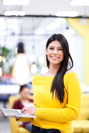 Beautiful girl with long dark hair working on a laptop in a modern office. Business colleagues in the backgorundの写真素材