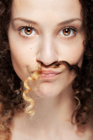 Beautiful brunette girl with long curly hair studio portrait. Lock of hair under the nose imitating moustache. Focus on the hair.の写真素材