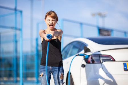 Beautiful young girl next to an electric car. Posing and holding a charging cable.の写真素材
