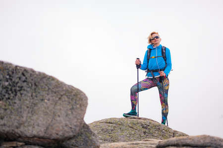 Hiking girl with poles and backpack standing on rocks. Windy autumn day. Travel and healthy lifestyle outdoors in fall seasonの写真素材