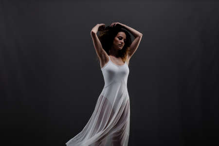 Girl with curly hair making ballet poses. Side lit silhouette of ballerina in white dress and black boots against black background.の写真素材