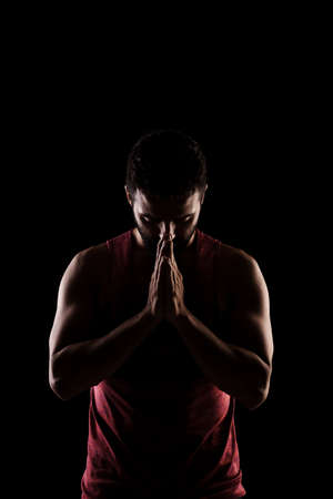 Side lit muscular Caucasian man silhouette. Athlete in red shirt praying against black background.の写真素材
