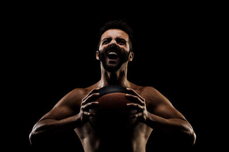 Basketball player holding a ball against black background. Side lit muscular Caucasian man silhouette.の写真素材