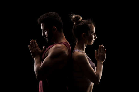 Fit couple posing together. Boy and girl praying back to back side lit silhouettes on black background.の写真素材