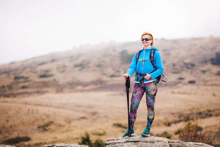 Hiking girl with poles and backpack standing on rocks. Windy autumn day. Travel and healthy lifestyle outdoors in fall seasonの写真素材
