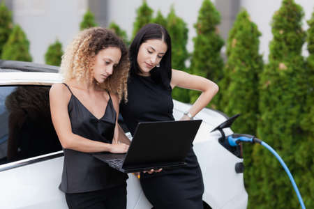 Two beautiful girls in black outfut smiling and posing. Businesswomen working on laptop while their electric car is charging in the background.の写真素材