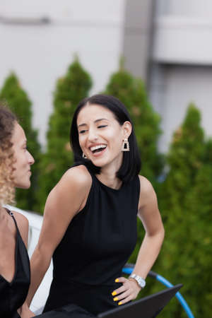 Two beautiful girls in black outfut smiling and posing. Businesswomen working on laptop while their electric car is charging in the background.の写真素材