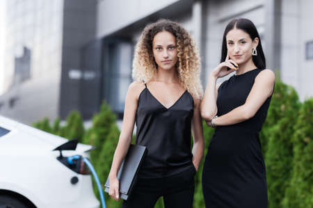 Two beautiful businesswomen in black outfut smiling and posing. Portrait of girls with laptop in front of electric car charging in the background.の写真素材
