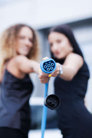 Two beautiful businesswomen in black outfut posing in front of an electric car. Girls holding blue charging cable. Shallow depth of field. Selective focus.の写真素材