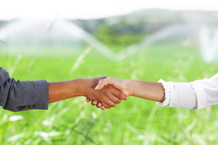 Business women shaking hands in a green field with agriculture irrigation system. Water sprinklers in the background.の写真素材