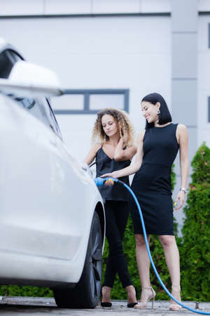 Two beautiful businesswomen in black outfut posing next to an electric car. Girls plugging blue cable in power charging socket.の写真素材
