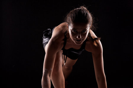 Side lit silhouette fit girl in race start position against dark background.