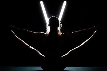 Fit woman practicing yoga poses. Silhouette girl doing exercise in studio against black background with v shaped white led tube light. No stress inner balance concept.の写真素材